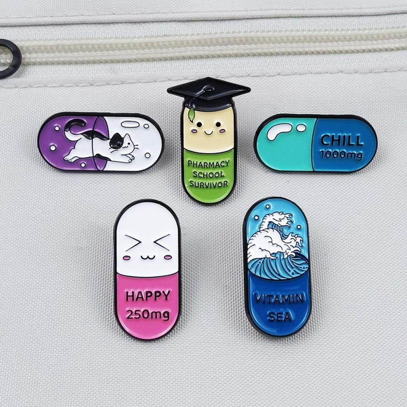 May include: A collection of enamel pins in the shape of pills. Designs include a cat, a graduate, and pills with the words "CHILL", "HAPPY", and "VITAMIN SEA". The pins are in various colors, including purple, blue, and pink.