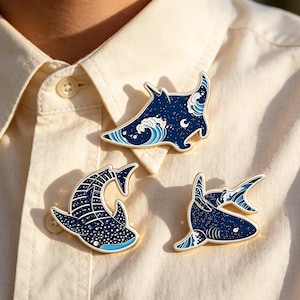 Puede incluir: Tres pines de esmalte con diseños de vida marina. Los pines incluyen una manta raya, un tiburón ballena y un tiburón, todos en tonos azules con detalles de olas y estrellas blancas. Los pines están sujetos a una camisa de color claro.