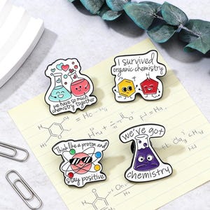 Könnte beinhalten: Vier Emaille-Anstecknadeln mit Designs zum Thema Chemie. Eine Anstecknadel zeigt zwei lächelnde Bechergläser mit dem Text "we have so much chemistry together". Eine andere sagt "I survived organic chemistry". Eine dritte Anstecknadel zeigt ein Atom mit Sonnenbrille und dem Text "Think like a proton and stay positive."