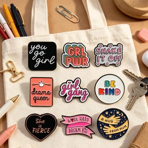 Pode incluir: Uma coleção de pins de esmalte com frases e designs motivacionais. Os pins incluem frases como "You go girl", "Girl gang" e "Be Kind". Os pins são de várias formas e cores, numa sacola branca.
