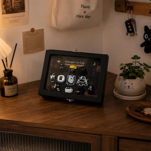 May include: A black display case with pins featuring cat-themed designs, including a coffee cup and a moon. The case is labeled "Midnight Cat Cafe." The display sits on a wooden surface, with a small potted plant and a decorative lamp nearby.