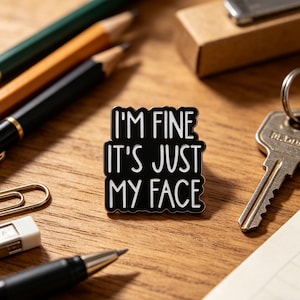 Op de afbeelding: Zwarte emaille pin met de tekst "I'M FINE IT'S JUST MY FACE" in het wit. De pin ligt op een houten oppervlak met potloden, een pen, een paperclip en een sleutel.