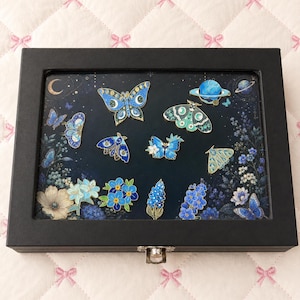 May include: A black rectangular display box containing a collection of enamel pins featuring butterflies, moths, flowers, and a planet. The pins are arranged against a dark blue background with floral and celestial motifs. Text reads: Includes display box+curated enamel pins.