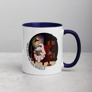Może przedstawiać: Biały ceramiczny kubek z granatowym wnętrzem i uchwytem. Kubek przedstawia kreskówkową ilustrację ducha czytającego książkę w przytulnym otoczeniu, z napisem "BELIEVE IN YOURSELF" otaczającym wzór.