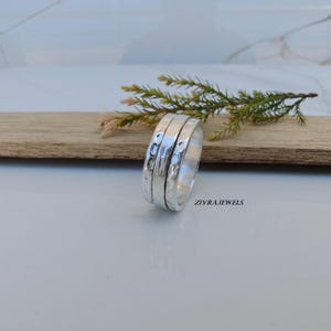 Puede incluir: Un anillo giratorio de plata con una banda central pulida flanqueada por dos bandas exteriores con acabado martillado y texturizado. El anillo tiene grabado el texto ZIVRAJEWELS en el lateral. El diseño consta de tres anillos metálicos unidos.