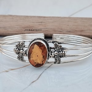 May include: A silver cuff bracelet featuring a central oval-cut amber-coloured gemstone surrounded by a twisted metal border. Two detailed bee motifs are positioned on either side of the stone, set against a multi-strand band. Text reads ZIVRAJEWELS.