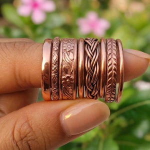 Könnte beinhalten: Ein Set aus sieben Kupferringen mit verschiedenen Texturen und Mustern, darunter ein glattes Band, ein geometrisches Rautenmuster, ein Blumenmotiv, ein schlichtes Band, eine geflochtene Seilstruktur, ein blattartiges Muster und ein schlichtes Band.