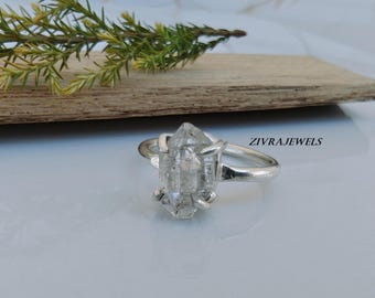 Può includere: Un anello in argento con un cristallo grezzo esagonale trasparente tenuto da quattro griffe. La fascia è in argento liscio e lucido. Il testo ZIVRAJEWELS è stampato in nero sul lato destro dell'anello. Il cristallo presenta inclusioni visibili.
