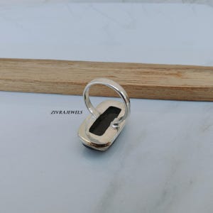 Pu&ograve; includere: Un anello d'argento con una pietra rettangolare di colore scuro. L'anello &egrave; incastonato in una fascia d'argento ed &egrave; esposto su una superficie chiara con un accento in legno. Il testo "ZIVRAJEWELS" &egrave; visibile.