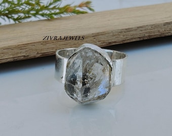 Può includere: Un anello in argento con una grande pietra di cristallo grezzo, trasparente e sfaccettata, incastonata in un'ampia fascia di metallo lucido. L'anello è posizionato davanti a un'asse di legno con il testo ZIVRAJEWELS stampato in nero.
