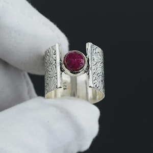 May include: A wide sterling silver ring featuring an intricate embossed scrollwork pattern on the band. The centre of the ring displays a round, deep pinkish-red gemstone set in a smooth bezel, with the hallmark 925 stamped on the inner band.
