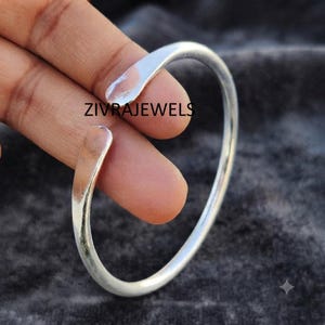 Puede incluir: Una pulsera de brazalete abierto en tono plateado con un acabado liso y pulido y extremos redondeados. La banda de metal presenta un diseño circular simple que es flexible para ajustar. El texto ZIVRAJEWELS es visible en el metal.