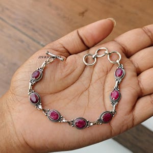 以下が含まれることがあります： 楕円形の深紅色の宝石をあしらったシルバーブレスレット。 ブレスレットにはトグルクラスプが付いており、相互接続されたリンクで構成されています。 宝石は精巧で詳細なベゼルにセットされています。 ブレスレットには「ZIVRA JEWELS」の文字が見えます。