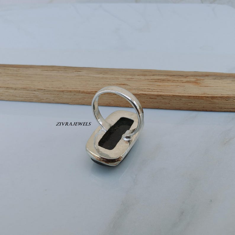 Pu&ograve; includere: Un anello d'argento con una pietra rettangolare di colore scuro. L'anello &egrave; incastonato in una fascia d'argento e viene esposto su una superficie chiara con un elemento in legno. Il testo "ZIVRAJEWELS" &egrave; visibile.