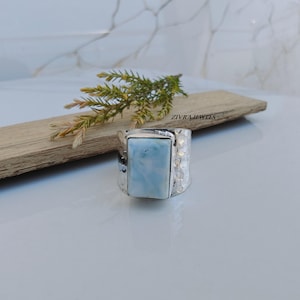 May include: A silver ring featuring a rectangular, light blue, marbled stone set in a hammered metal band. The ring is positioned near a small green sprig, and the text ZIVRA JEWELS is visible on the wooden surface behind the jewelry piece.