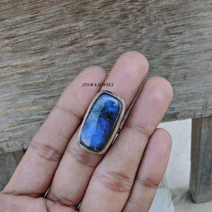 Pu&ograve; includere: Un anello con una pietra preziosa di labradorite rettangolare incastonata in una fascia d'argento lucido. La pietra mostra un profondo riflesso blu iridescente con inclusioni scure. Il testo ZIVRAJEWELS &egrave; visibile sopra l'anello.