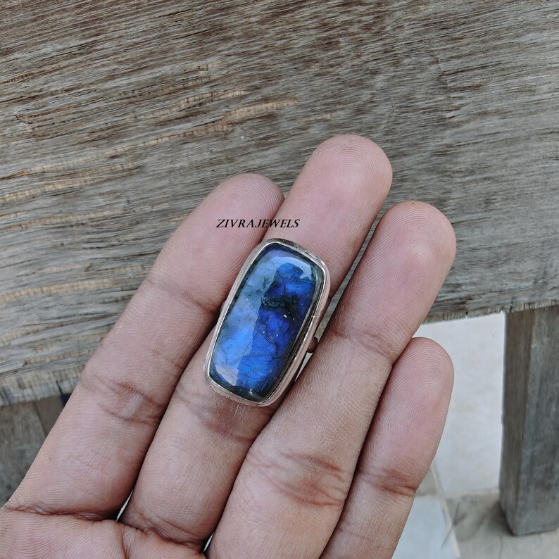 Pu&ograve; includere: Un anello con una pietra preziosa di labradorite rettangolare incastonata in una fascia d'argento lucido. La pietra mostra un profondo riflesso blu iridescente con inclusioni scure. Il testo ZIVRAJEWELS &egrave; visibile sopra l'anello.