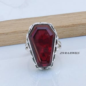 May include: A silver ring featuring a large, faceted, deep red gemstone cut into a coffin shape. The metal setting has a textured, ornate border surrounding the stone. The text ZIVRAJEWELS is printed in black on the right side of the ring.