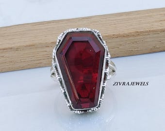 Può includere: Un anello in argento con una grande pietra preziosa rosso scuro sfaccettata, tagliata a forma di bara. La montatura in metallo presenta un bordo testurizzato e decorato attorno alla pietra. Il testo ZIVRAJEWELS è stampato sul lato destro.