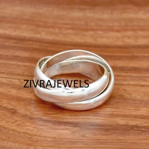Pode incluir: Um anel de cor prateada composto por três bandas entrelaçadas que se sobrepõem para formar uma estrutura circular única. A superfície metálica é lisa e reflexiva, com o texto ZIVRAJEWELS impresso no centro do anel.
