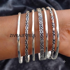 Peut inclure: Un ensemble de six bracelets rigides en métal argenté présentant divers motifs, notamment des bandes lisses, un motif ondulé, une décoration florale et une texture perlée. Le texte ZIVRAJEWELS est visible sur les bracelets portés ensemble.