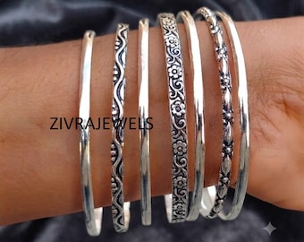 Può includere: Un set di sei bracciali rigidi in metallo color argento con vari design, tra cui fasce lisce, un motivo ondulato, un decoro floreale e una trama a perline. Il testo ZIVRAJEWELS è visibile sui bracciali indossati tutti insieme.
