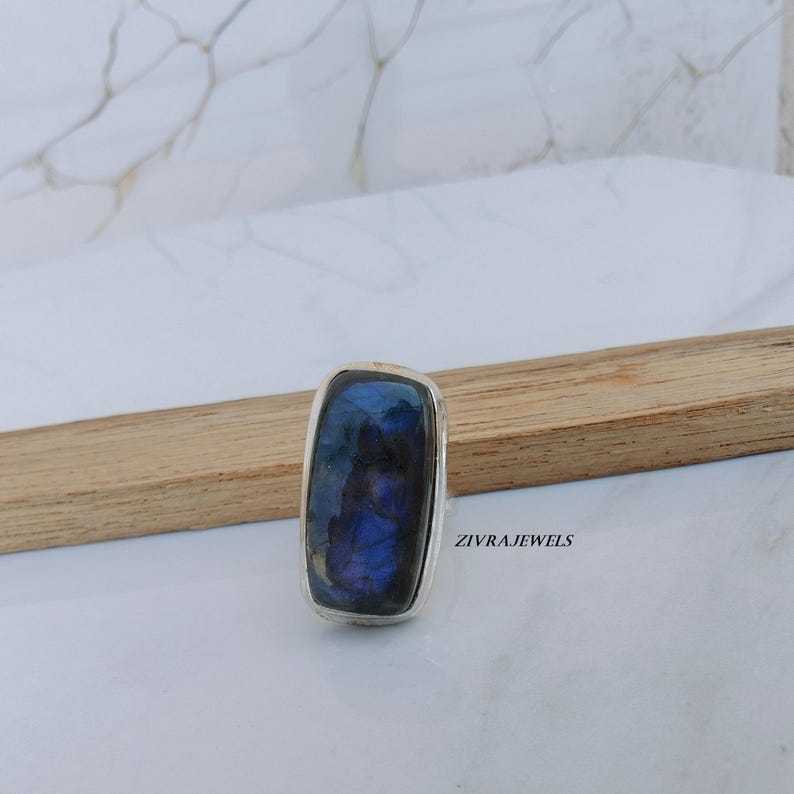 Pu&ograve; includere: Un anello rettangolare con una grande pietra preziosa blu iridescente incastonata in una fascia d'argento. La gemma mostra sfumature di blu e nero. L'anello &egrave; esposto su una superficie chiara con un accento in legno. Il testo "ZIVRAJEWELS" &egrave; visibile.