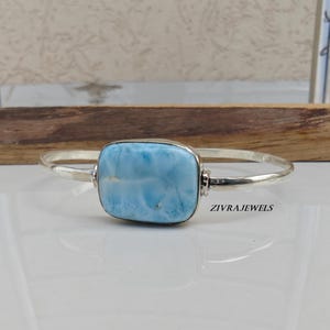 May include: A silver bracelet featuring a rectangular, light blue larimar stone. The stone has a marbled appearance with white and blue hues. The bracelet is displayed on a white surface.