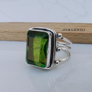 May include: A silver ring featuring a large, rectangular, light green gemstone. The ring has a triple-band design and small decorative spheres. The text "ZIVRAJEWELS" is visible on the wooden background.