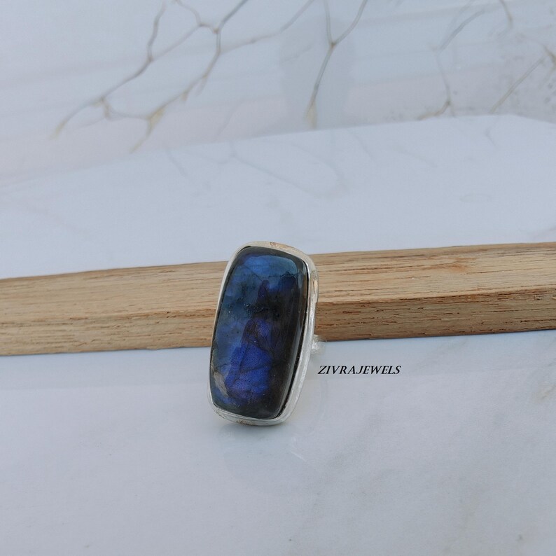 Pu&ograve; includere: Anello rettangolare in argento con una grande pietra di labradorite iridescente. La pietra mostra sfumature di blu e viola. L'anello &egrave; su una superficie chiara con un elemento in legno. Il testo "ZIVRAJEWELS" &egrave; visibile.