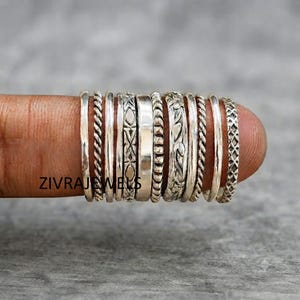 Könnte beinhalten: Ein Set aus elf silberfarbenen Ringen mit verschiedenen Texturen und Mustern, darunter gedrehte Kordeln, geometrische Formen, glatte Bänder und komplizierte florale Gravuren, die nebeneinander auf einem Finger angeordnet sind.