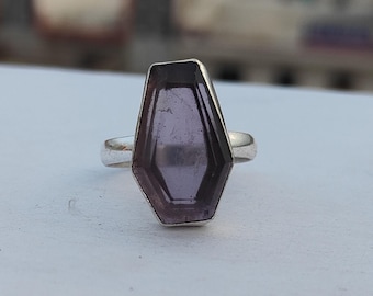 Può includere: Un anello in argento con una grande pietra preziosa viola sfaccettata a forma di bara, incastonata in una semplice fascia metallica. La pietra ha un aspetto traslucido con inclusioni interne visibili e una superficie piatta lucida.