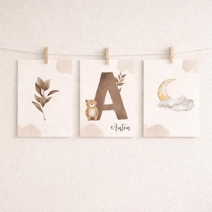 Puede incluir: Tres impresiones artísticas en acuarela colgadas de una cuerda. Las impresiones presentan una hoja, la letra "A" con un osito de peluche y el nombre "Anton", y una luna creciente con nubes. Las impresiones son en tonos marrones y beige.