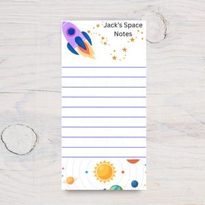 May include: A rectangular notepad featuring a purple rocket ship, gold stars, and the text Jack's Space Notes at the top. The center has blue horizontal lines for writing, and the bottom displays a solar system graphic with a yellow sun and planets.