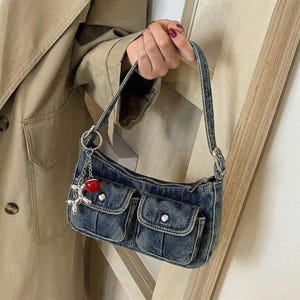 Könnte beinhalten: Eine blaue Jeans-Umhängetasche mit zwei Vordertaschen und einem silbernen Reißverschluss. Die Tasche hat einen silbernen Anhänger mit einer roten Kirsche und einem hundeformigen Anhänger. Die Tasche wird von einer Person gehalten, die einen beigen Trenchcoat trägt.