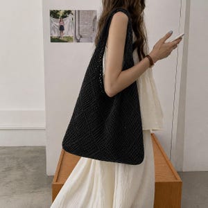Puede incluir: Un bolso tote negro de tejido abierto con un patrón de diamantes. El bolso se lleva al hombro. El bolso está hecho de un material tejido. El bolso es de color neutro y se puede usar para ir de compras o llevar objetos personales.