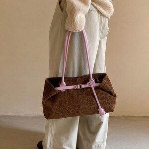 May include: A brown suede shoulder bag featuring a subtle leopard print pattern. The bag includes two long, light pink shoulder straps and a matching light pink horizontal strap across the front, secured with a silver-toned metal buckle.