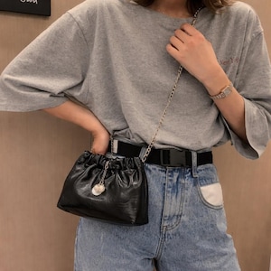 Puede incluir: Un pequeño bolso de mano de cuero negro fruncido con una correa de cadena dorada. El bolso tiene un pequeño dije redondo. La persona lleva una camiseta gris y vaqueros azules.