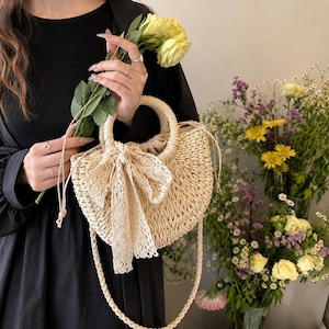 May include: A woven straw handbag with a rounded shape and a cream-colored lace bow. The bag has a braided handle and a long, braided strap. The bag is held by a person wearing a black dress, and there are yellow roses and other flowers in the background.