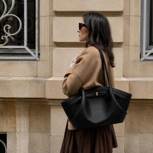 May include: A black leather tote bag with a pebbled texture, featuring dual shoulder straps, a curved silhouette, and a small gold-toned metal accent on the front. The bag is carried on the shoulder and has a structured, wide base design.