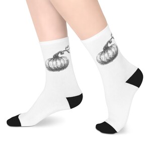 May include: A pair of white crew socks featuring a black ink-style sketch of a pumpkin on the outer side of each sock. The socks have solid black heels and black toe sections, providing a clear contrast against the white fabric of the socks.