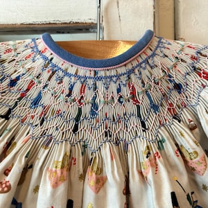 May include: A white smocked dress with a blue collar and a festive print of marching toy soldiers and presents. The smocking is done in pink, blue, and green thread.