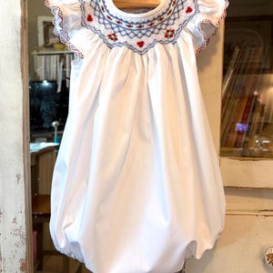 May include: White baby bubble dress with a smocked bodice. The bodice features blue and red embroidery with a scalloped edge. The dress has short, ruffled sleeves and a full, gathered skirt. The dress is hanging on a wooden hanger.