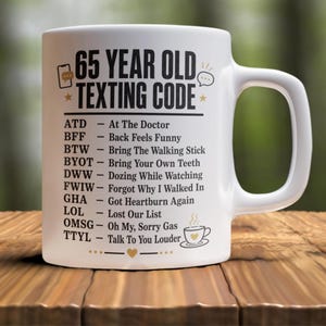 May include: A white ceramic mug featuring black text that reads 65 YEAR OLD TEXTING CODE, followed by a list of humorous acronym definitions like ATD for At The Doctor and BYOT for Bring Your Own Teeth, accented with small gold icons of a phone and heart.