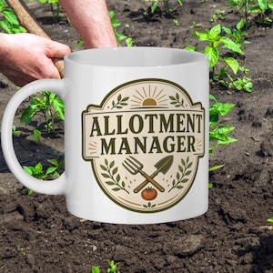 May include: White ceramic mug featuring the text "ALLOTMENT MANAGER" in a brown and green design. The design includes a fork, shovel, and tomato. The mug is in a garden setting.