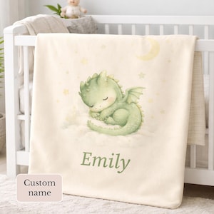 Puede incluir: Una manta de bebé color crema con una ilustración de acuarela de un dragón verde durmiendo sobre una nube. La manta tiene un nombre personalizado, "Emily", en escritura verde. También se representan una luna y estrellas.