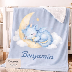 May include: A light blue baby blanket featuring a sleeping blue dragon on a yellow crescent moon. The blanket has a white sherpa lining and the name "Benjamin" is printed on the front. The words "Custom name" are printed on a small, light-colored rectangle.