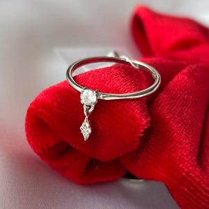 May include: A silver ring with a dangling diamond-shaped charm and a round diamond. The ring is set against a red velvet background and a white satin surface.