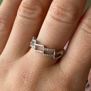 May include: A silver ring with a geometric design featuring rectangular links. The ring is adorned with small, sparkling stones, adding a touch of elegance. The ring is shown on a hand.