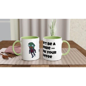 May include: Two white ceramic coffee mugs with green handles and rims. One mug features a cartoon zombie illustration, the other has the text "Don't be a zombie - drink your coffee."